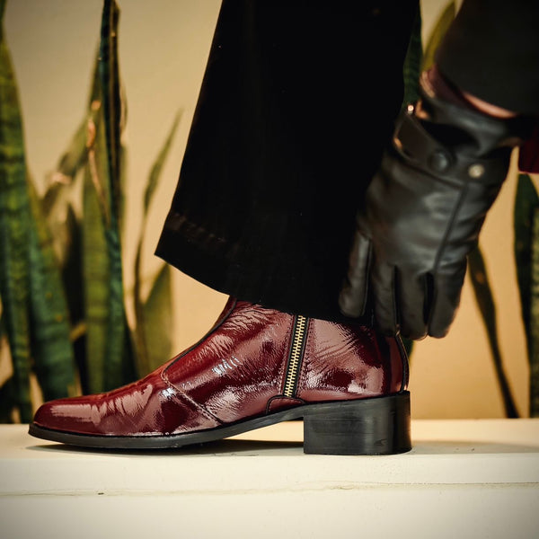 A close-up of a stylish cherry red pointed-toe boot in glossy patent leather with a side zip closure, worn by a person dressed in black trousers and leather gloves. The boot is elevated on a white platform with green snake plants in the background, creating a bold and luxurious contrast.