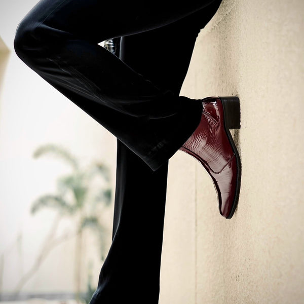 Men’s luxury cherry red pointed-toe Chelsea boots in patent leather with side zip closure, worn with black trousers and leather gloves; sleek and stylish footwear ideal for formal and high-fashion outfits.