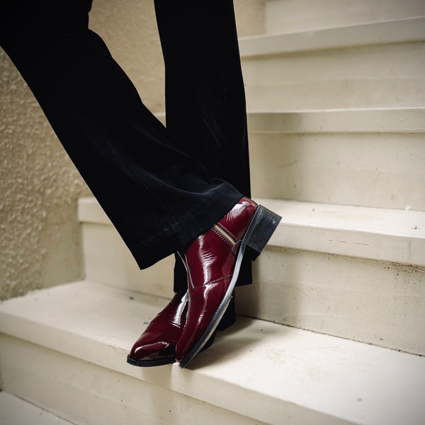 Men’s luxury cherry red pointed-toe Chelsea boots in patent leather with side zip closure, worn with black trousers and leather gloves; sleek and stylish footwear ideal for formal and high-fashion outfits.